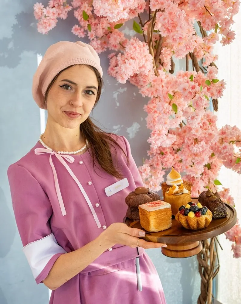 Viva La Dough baker in pink uniform holding a tray of pastries under cherry blossoms